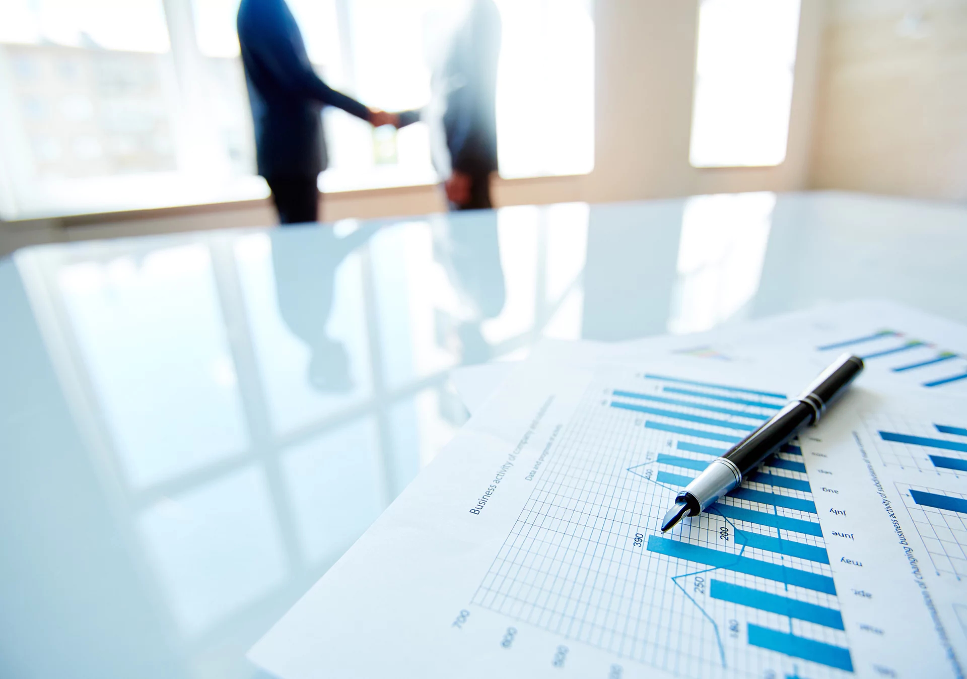 Business meeting handshake in bright office with financial charts and pen on reflective table, representing corporate transparency and compliance