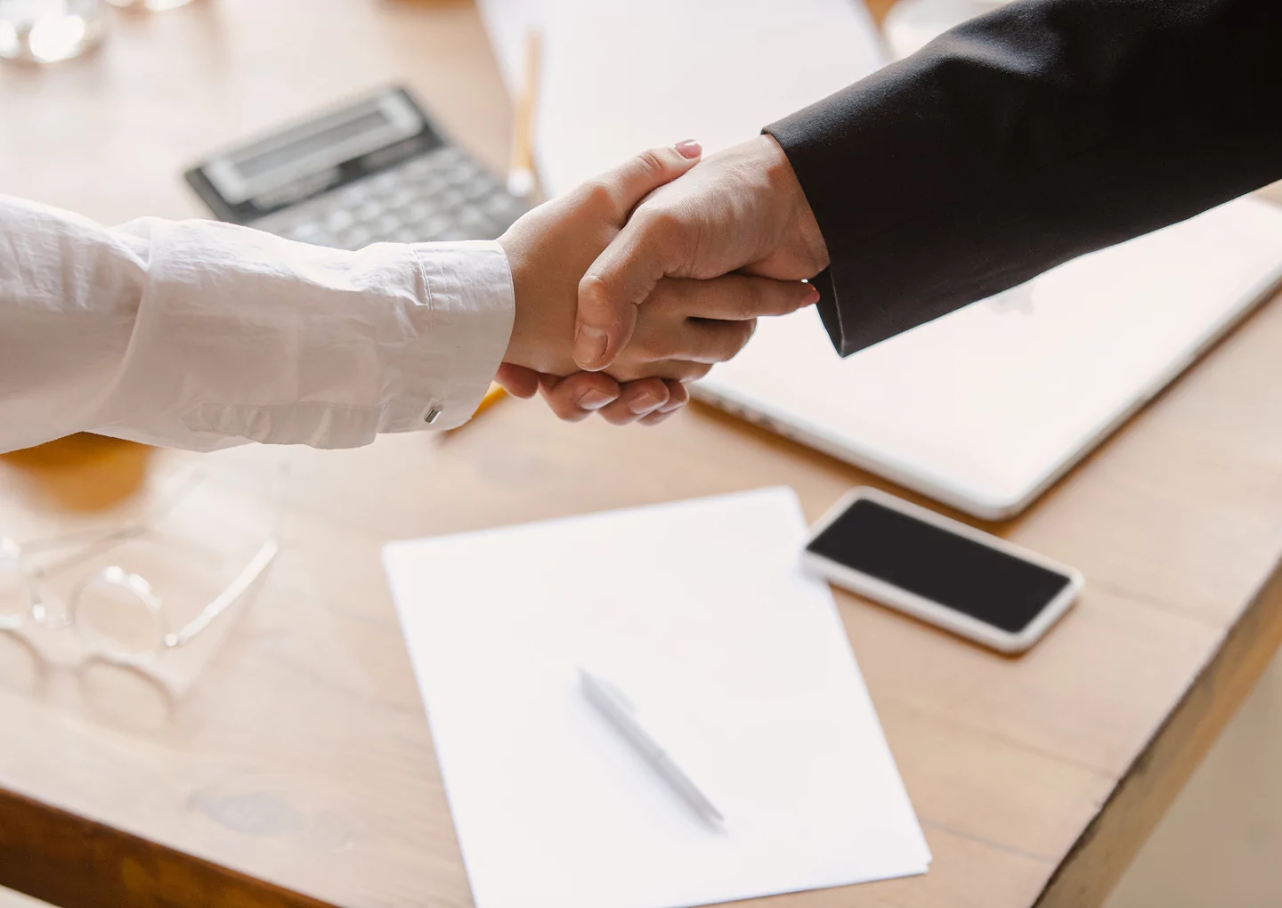 Business professionals shaking hands over a desk with documents, a smartphone, and a laptop, symbolizing partnership and R&D collaboration.