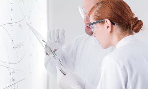 Scientist's hands writing on a clipboard surrounded by lab equipment including petri dishes, test tubes with blue and pink liquids, and a dropper on a white laboratory table